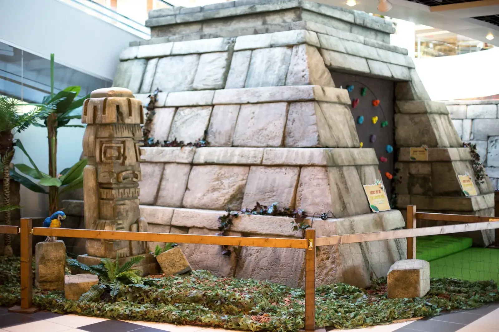 Pyramide en pierre reconstituée avec mur d'escalade, totem sculpté et végétation, décor installé dans une galerie commerciale